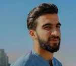 A close-up outdoor portrait of a young man with facial hair, exuding confidence under a clear sky.
